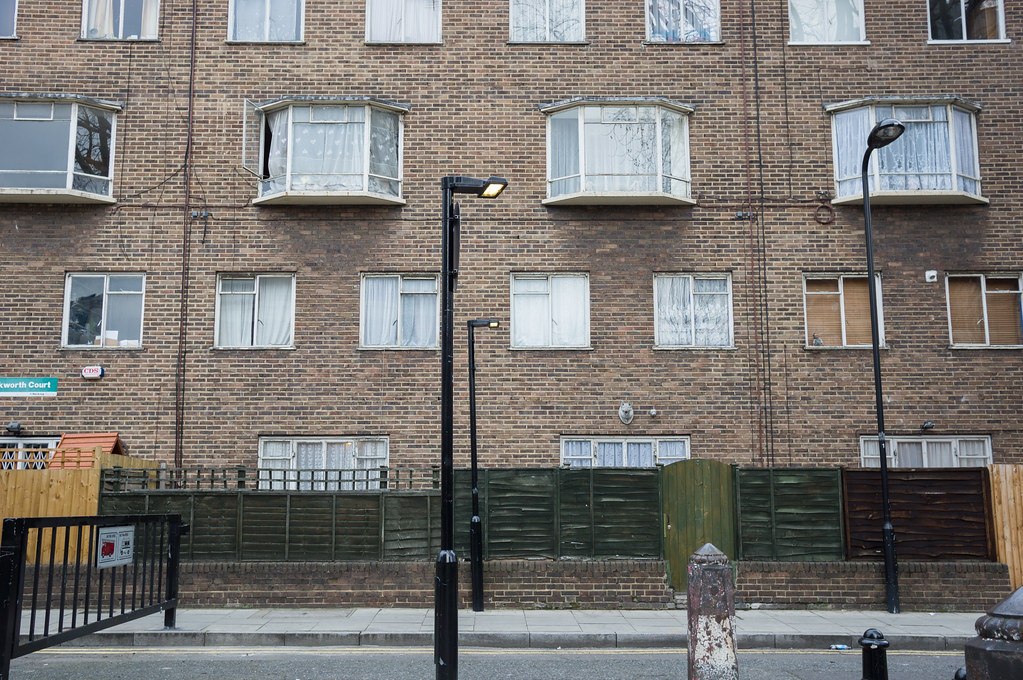 A near-symmetrical view of a London brick council estate facade, raking late-afternoon light, three identical streetlamps lined up across the frame casting precise shadows. Bay windows with translucent net curtains are arranged in two storeys; a green-painted wooden fence runs along a basement-level area at the bottom; the pavement and a black metal handrail are visible in the foreground. There are no figures, no signs, no incident.